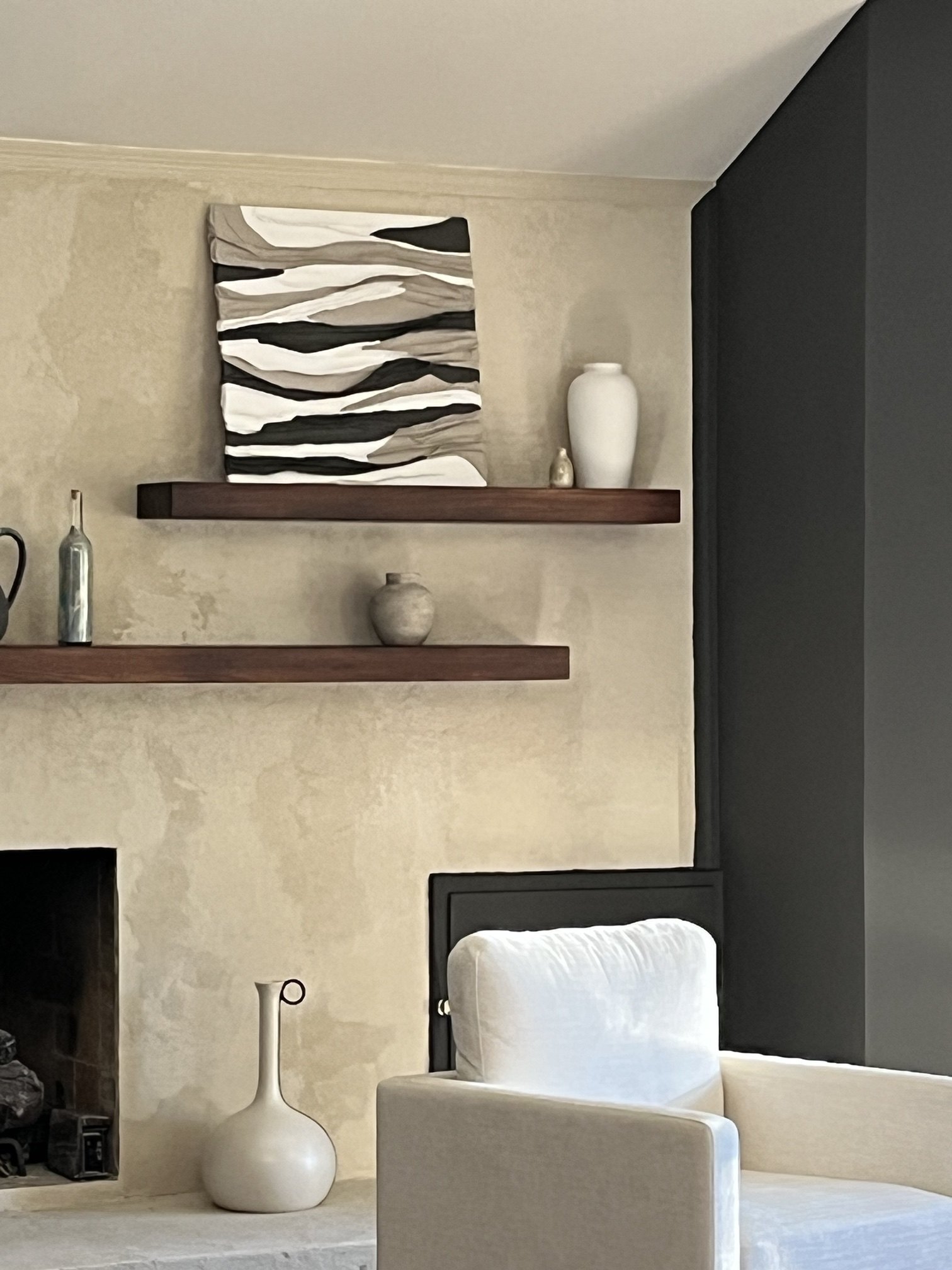 A square textured piece of layered folds in white, taupe and charcoal, styled on a walnut shelf above a plaster fireplace with cream vases