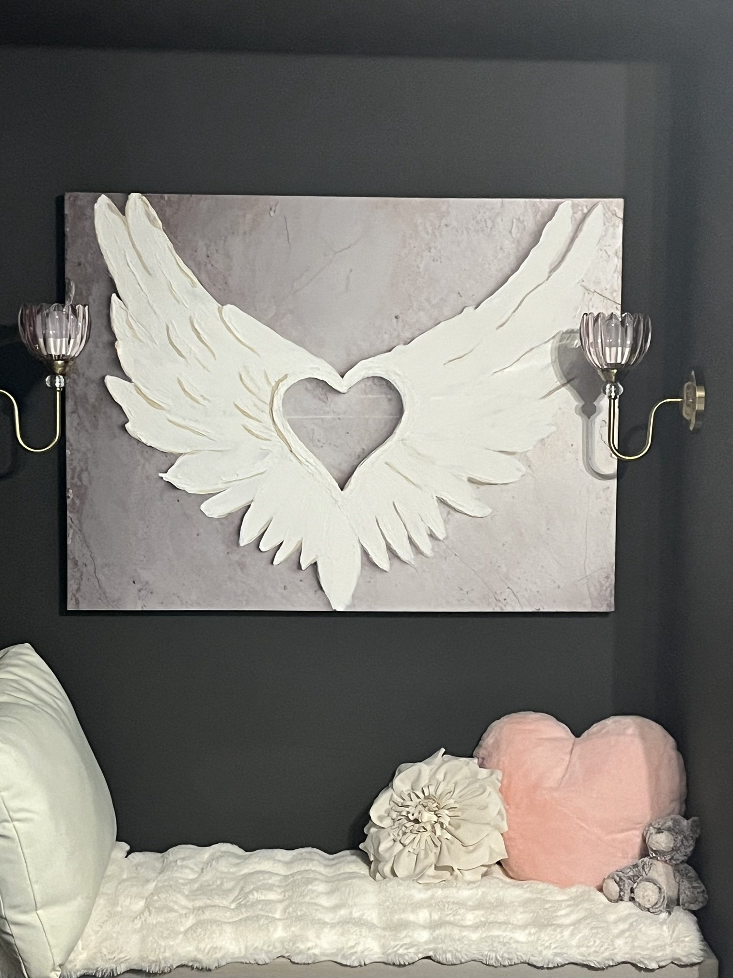 Sculpted white angel wings framing an open heart, on a soft blush-grey plaster background, hung between brass sconces
