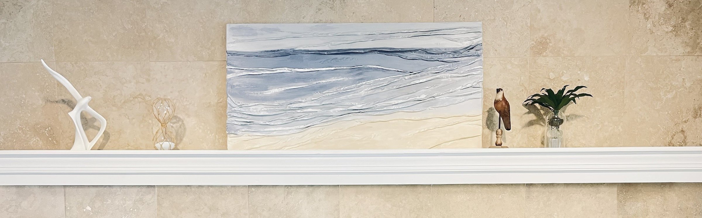 A wide textured piece in soft blue and cream, with layered folds like gentle waves meeting sand, displayed on a white mantel against travertine stone