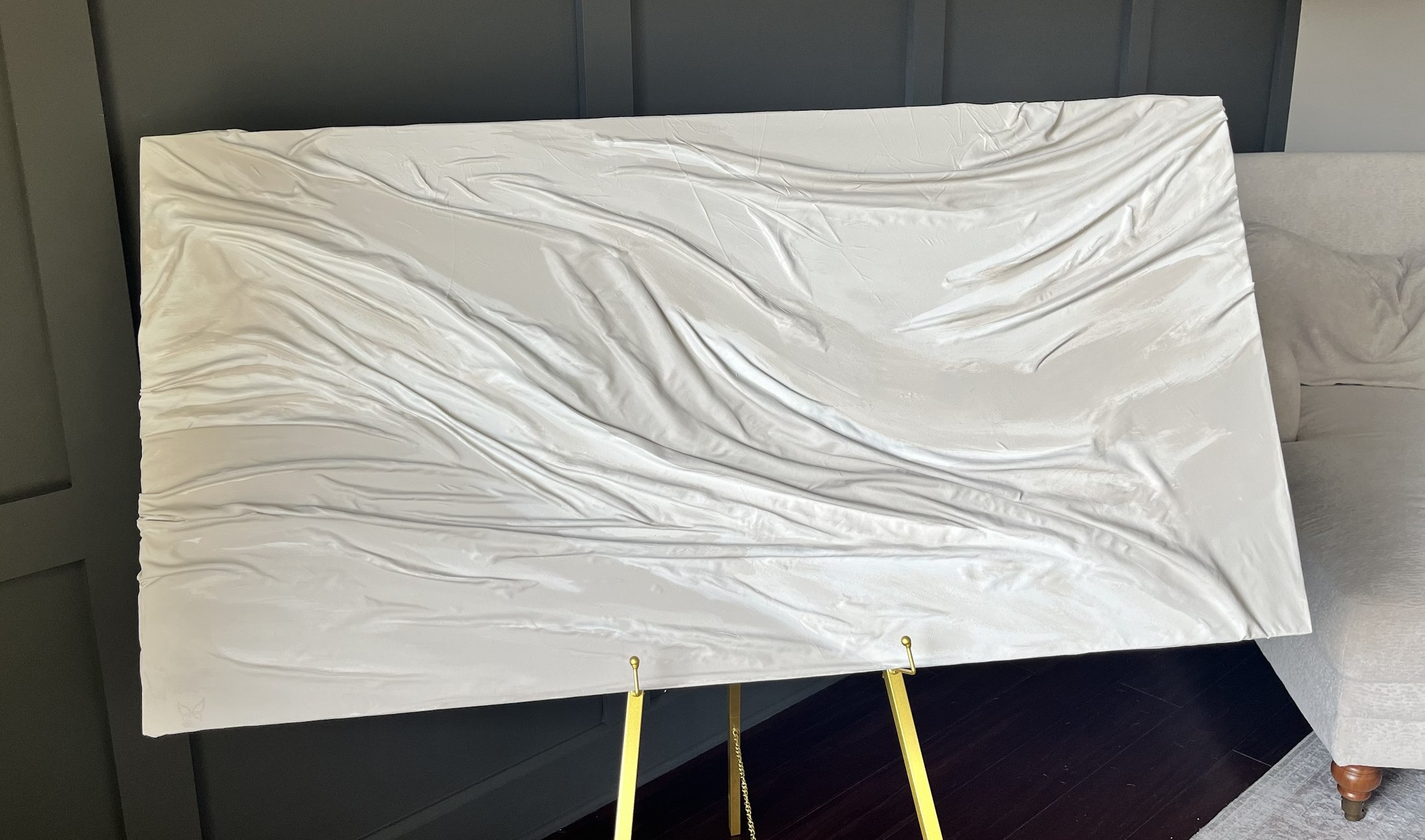 A wide cream textured piece of draped folds sweeping diagonally like wind across sand, on a gold easel against a charcoal paneled wall beside a soft upholstered sofa